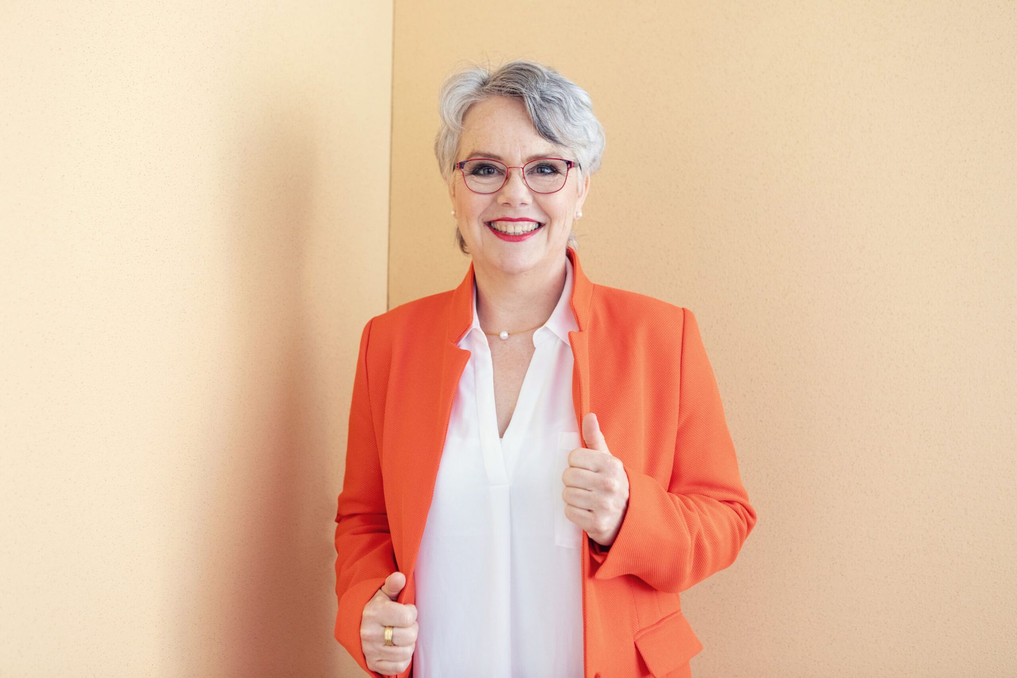 Imke Götz im orangefarbenen Blazer vor warmer Wand